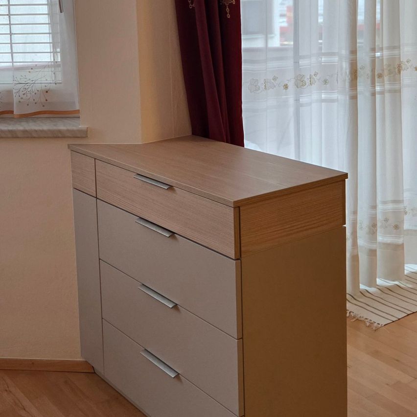 Ein hölzerner Kommode mit vier Schubladen steht in einem Raum mit einem Fenster, das von weißen Vorhängen bedeckt ist, und einem roten Vorhang im Hintergrund. Der Boden ist aus Holz.