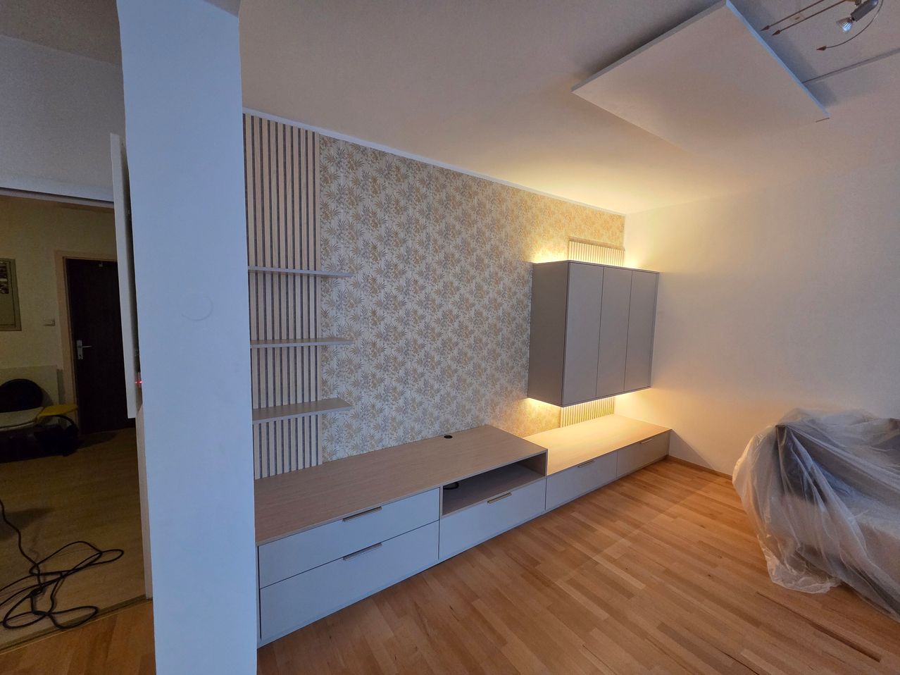 Ein leerer Raum mit Holzboden, einer Wand mit Blumentapete, einem Holzregal und Schränken auf der rechten Seite. Der Raum hat ein modernes Design.