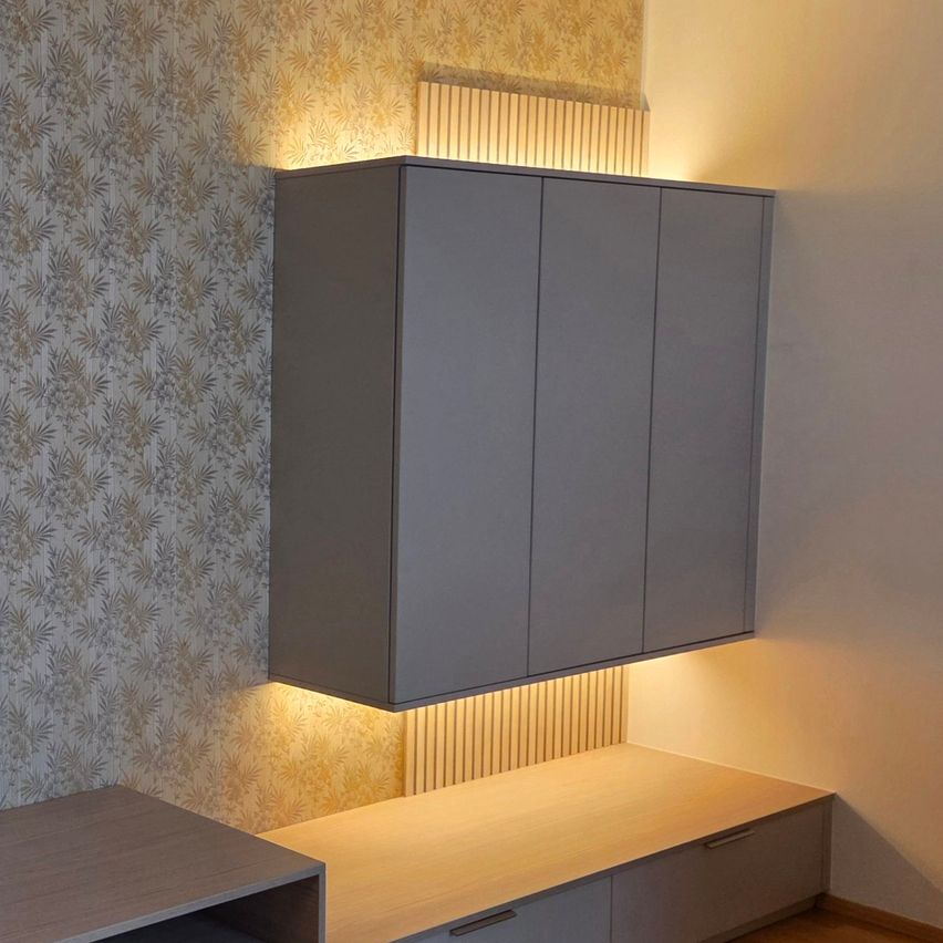 Ein moderner Raum mit einem grauen Schrank an der Wand, beleuchtet von gelben Lichtern. Unter dem Schrank befindet sich ein hölzerner Schreibtisch mit Schubladen. Die Wand hat eine Tapete mit Blattdesign.