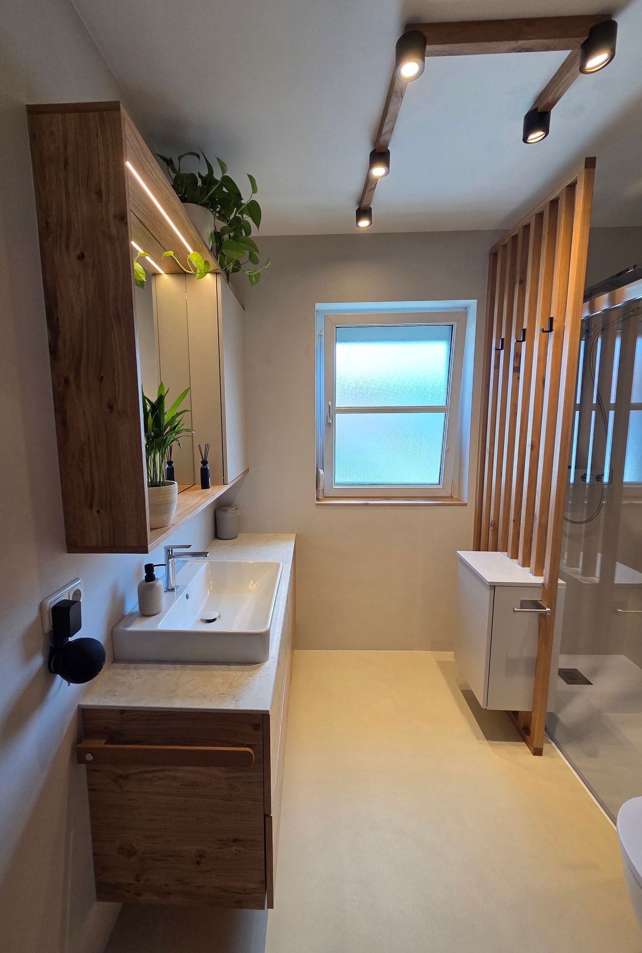 Ein modernes Badezimmer mit einem weißen Waschbecken, Holzschränken und einer Glasdusche. Auf dem Schrank stehen Topfpflanzen und ein Fenster mit Blick nach draußen.