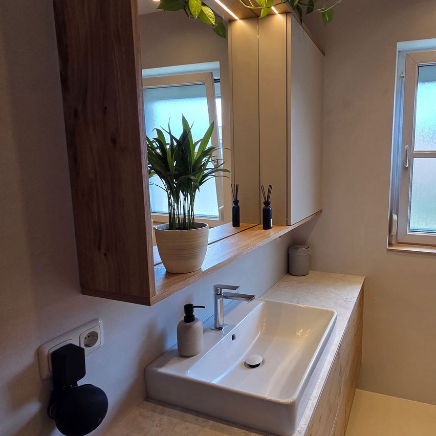 Ein modernes Badezimmer mit einem Holzschrank, einem Spiegel, einem weißen Waschbecken und einem Topf mit Pflanzen. Das Waschbecken hat einen Wasserhahn und einen Seifenspender. Der Schrank hat ein Regal mit einem Topf mit Pflanzen und zwei schwarzen Flaschen. Die Wand hat eine Steckdose und ein Fenster.
