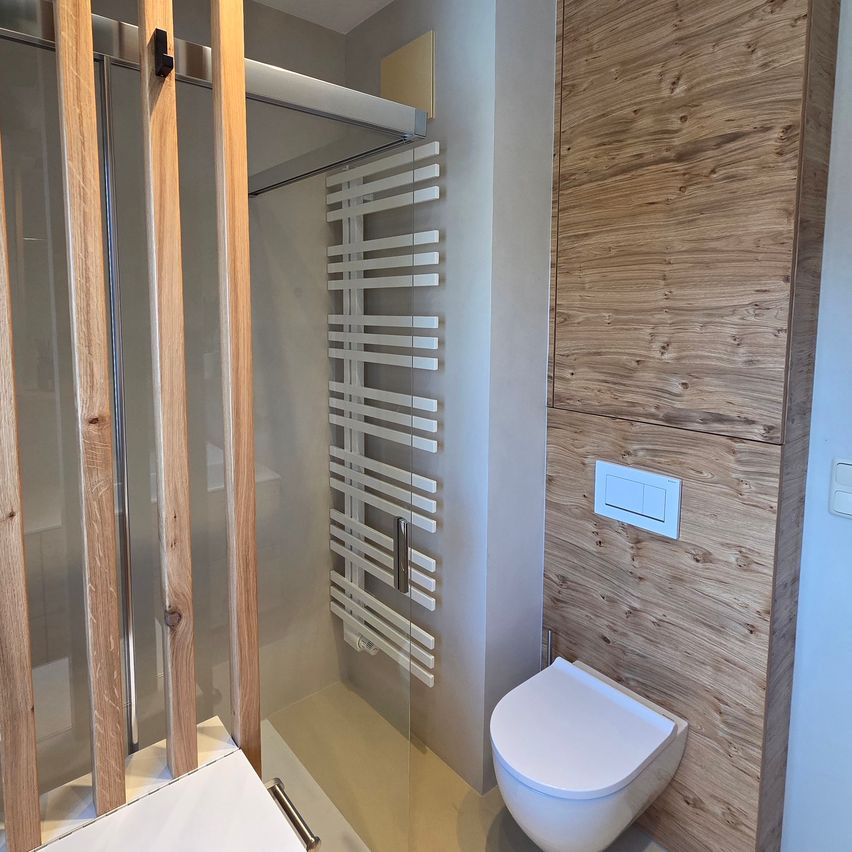 Ein modernes Badezimmer mit Holzwänden, einer Glasdusche und einem weißen Toilettensitz. Ein weißer Handtuchhalter ist an der Wand montiert.