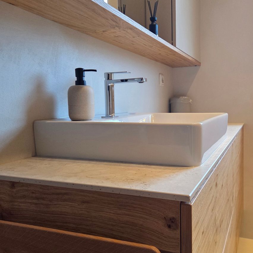 Ein modernes Badezimmer mit einem weißen Waschbecken, einem Holzschrank und einem Holzregal mit Spiegel und einer blauen Vase.