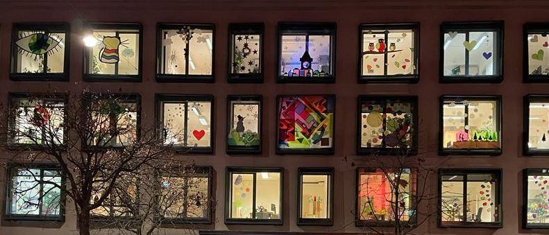 Ein Gebäude mit vielen Fenstern, die jeweils farbenfrohe Kunstwerke zeigen, steht in einer leeren Straße. Vor dem Gebäude stehen zwei Bäume und ein Fahrrad ist geparkt.