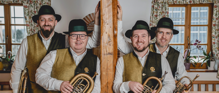 Four men in traditional Bavarian attire, including hats and vests, hold brass instruments and smile for the camera. They are positioned around a wooden post.