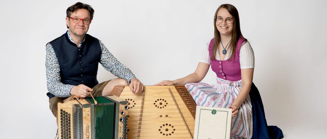 A man and a woman are sitting next to each other with traditional musical instruments. The man holds an accordion and the woman a zither. Both are smiling, and a book titled Lisa is between them.