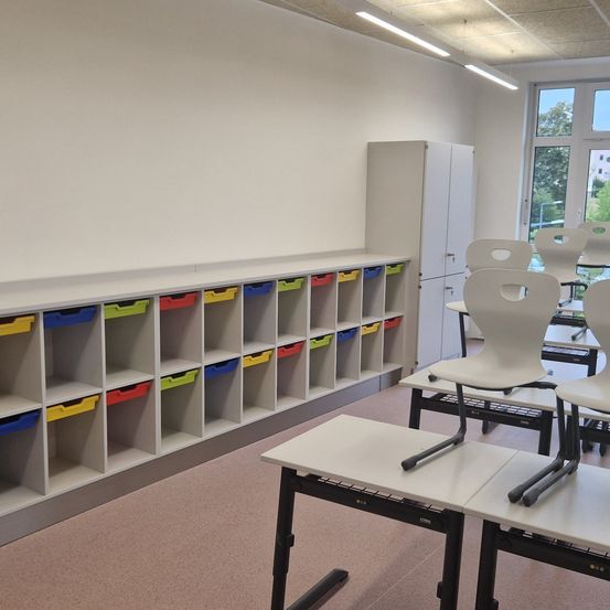 Ein leerer Klassenraum mit weißen Wänden und mehreren Regalen mit Fächern in verschiedenen Farben. Mehrere weiße Stühle stehen vor Tischen.