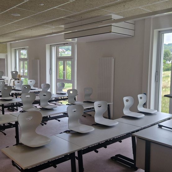 Ein leerer Klassenraum mit weißen Stühlen, Tischen und einem Heizkörper an der Wand. Der Raum hat große Fenster und eine Klimaanlage an der Decke.