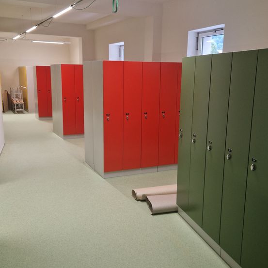 Ein Flur mit weißen Böden und Wänden zeigt eine Reihe roter und grüner Spinde. Links ist ein weißer Schrank zu sehen. Zwei Fenster an der Wand lassen natürliches Licht herein.