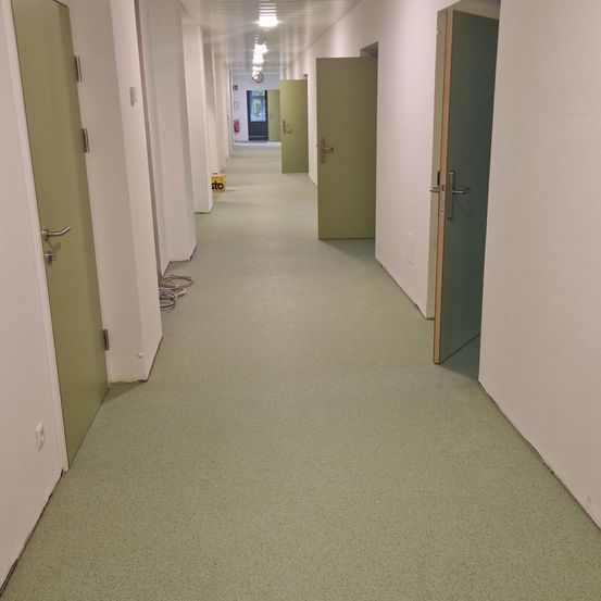 Ein leerer Korridor mit grünen Türen auf beiden Seiten. Der Boden ist grün und glatt. In der Ferne befinden sich zwei Feuerlöscher.