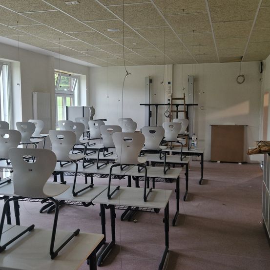 Ein leerer Klassenraum mit weißen Tischen und Stühlen, die in Reihen angeordnet sind, beleuchtet durch natürliches Licht aus Fenstern an einer Wand. Der Raum verfügt über eine weiße Decke mit einem Rastermuster, einem Heizkörper und einer braunen Aufbewahrungsbox.