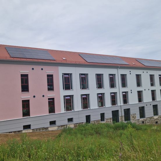 Ein mehrstöckiges Gebäude mit einem roten Dach, das mehrere Solarpanels aufweist, ist von hohem Gras umgeben, unter einem bewölkten Himmel.