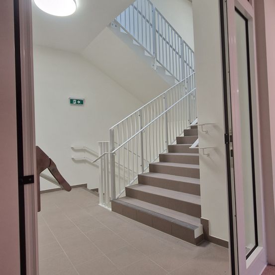 Ein Innenansicht eines Gebäudes mit gefliestem Boden, weißen Wänden und einer Treppe mit Handläufen. Das Ausfahrtsschild ist über der Treppe an der Wand montiert.