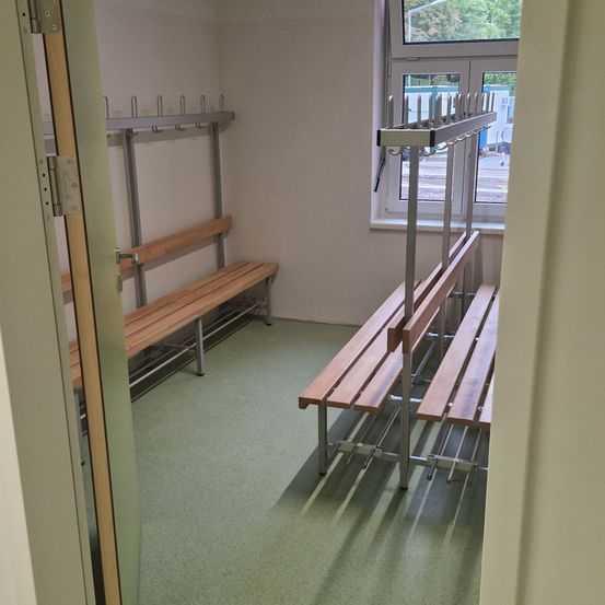Ein leerer Umkleideraum mit Holzbänken und Metallhaken an den Wänden, gesehen durch eine offene Tür. Ein Fenster zeigt die Ansicht einer Person, die draußen läuft.