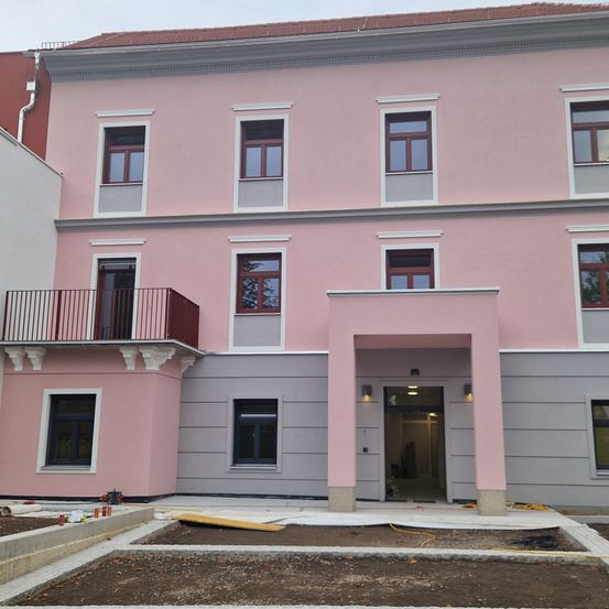 Das rosa Gebäude mit grauen Akzenten und großen Fenstern wird gebaut. Ein Balkon mit Geländer befindet sich im zweiten Stock.