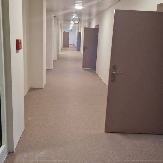 Ein leerer Flur mit weißen Wänden und einem braunen Boden. Mehrere Türen sind geschlossen, und ein Feuerlöscher ist an der linken Wand angebracht.