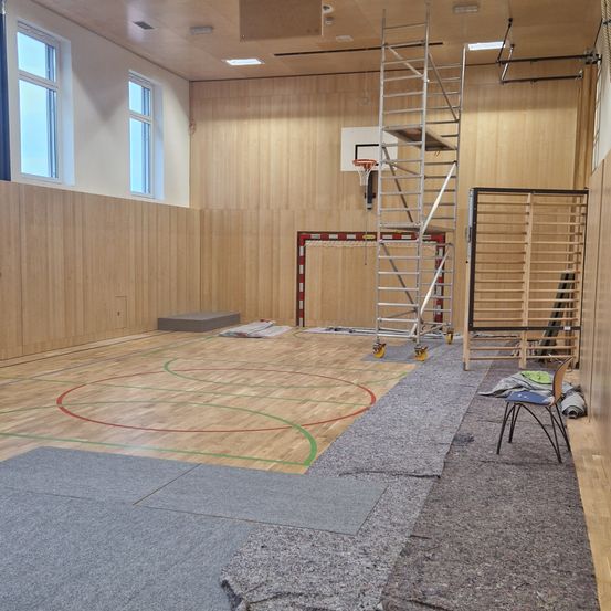 Eine leere Indoor-Sporthalle mit Holzwänden, einem Basketballkorb, einer Leiter und einem kleinen Stuhl. Der Boden hat Basketballmarkierungen.