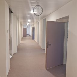Ein leerer Flur mit geschlossenen Türen, einer Uhr, die 10:11 anzeigt, und Teppichböden.