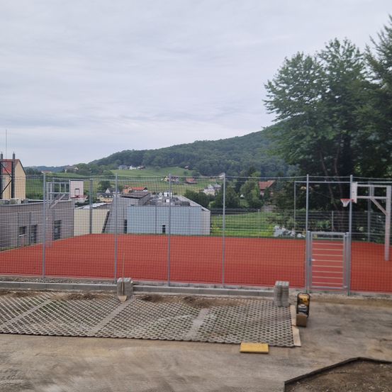 Ein Outdoor-Basketballplatz mit roter Oberfläche und Metallzaun befindet sich im Bau. Bäume und Häuser sind im Hintergrund sichtbar.
