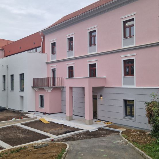 Ein zweistöckiges Gebäude mit rosa Außenwänden und grauen Akzenten, einschließlich eines Balkons und einer Einfahrt, im Bau mit einem teilweise gepflasterten Bereich und Grünflächen.