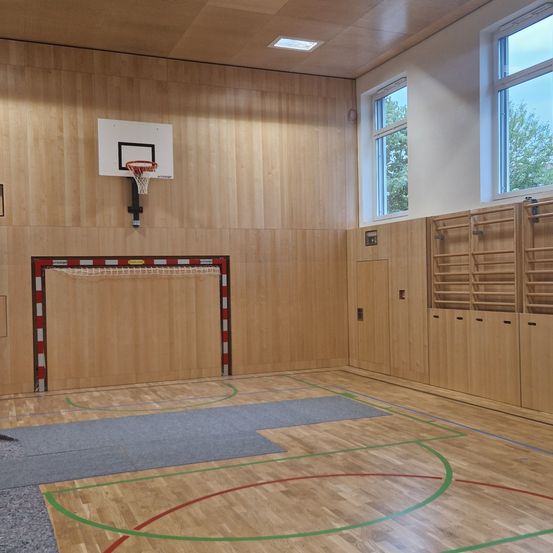 Eine Indoor-Sportanlage mit Holzwänden und -boden, einem Basketballkorb, einem rot-weiß-roten Tor und zwei Fenstern.