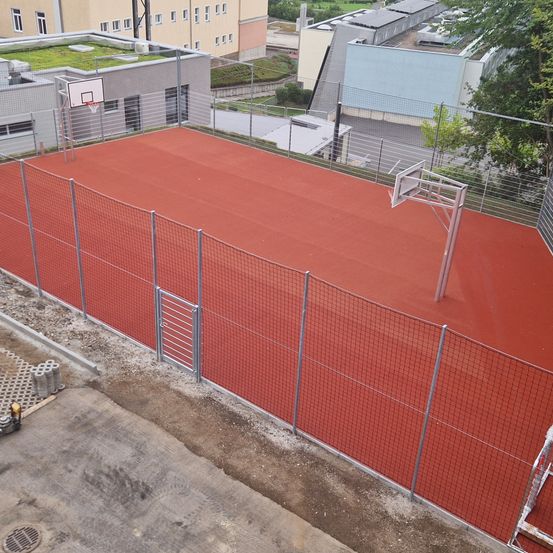 Ein Luftbild eines Basketballplatzes, umgeben von einem Zaun, mit einem roten Boden und Metallkörben auf beiden Seiten. Bäume und Gebäude sind im Hintergrund zu sehen.