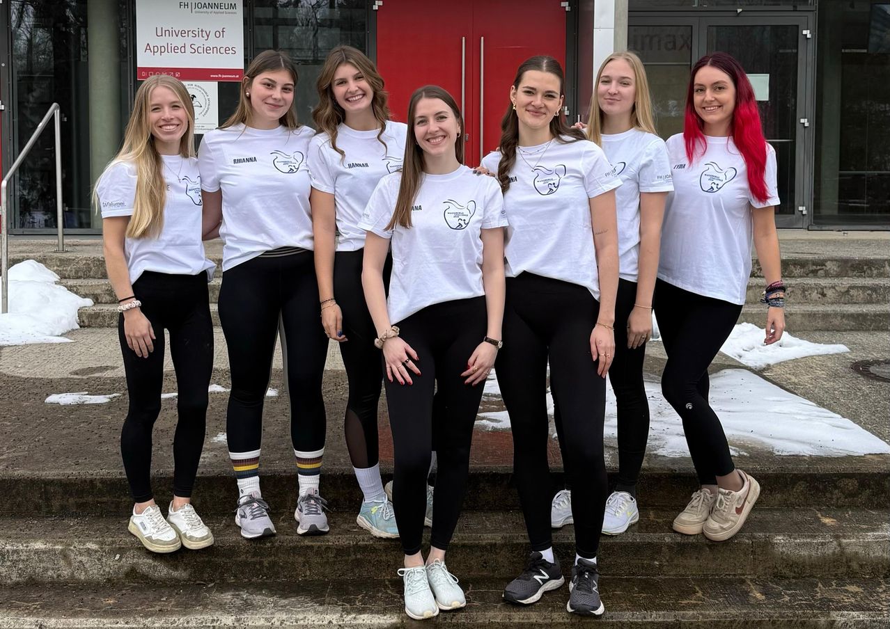 Sieben Frauen stehen auf Stufen vor einem Gebäude mit einer roten Tür und tragen weiße T-Shirts und schwarze Leggings. Sie lächeln und scheinen für ein Foto zu posieren.