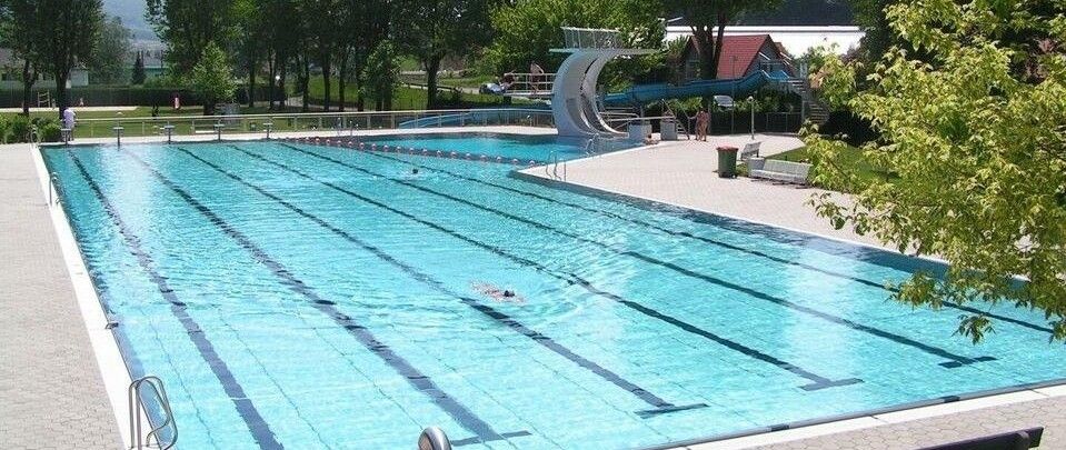 Ein Freibad mit Sprungturm und einer blauen Rutsche. Bäume und Häuser sind im Hintergrund. Eine Person schwimmt.