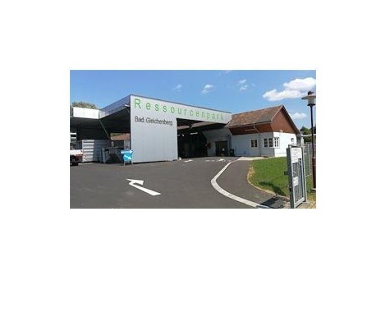 Ein Bild zeigt ein Gebäude mit dem Namen 'Ressourcenpark'. Es hat eine große weiße Wand und ein braunes Dach. Der Eingang befindet sich auf der linken Seite. Ein blauer Kasten steht auf dem Boden. Eine Straßenlaterne befindet sich auf der rechten Seite.