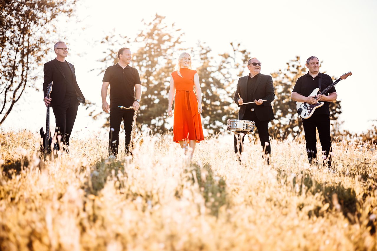 Eine Band aus fünf Musikern steht in einem Feld mit hohem Gras, jeder hält sein Instrument. Die Frau in der Mitte trägt ein oranges Kleid. Der Mann links hält eine Saxophon. Der Mann rechts hält einen Drumstick und eine Gitarre.
