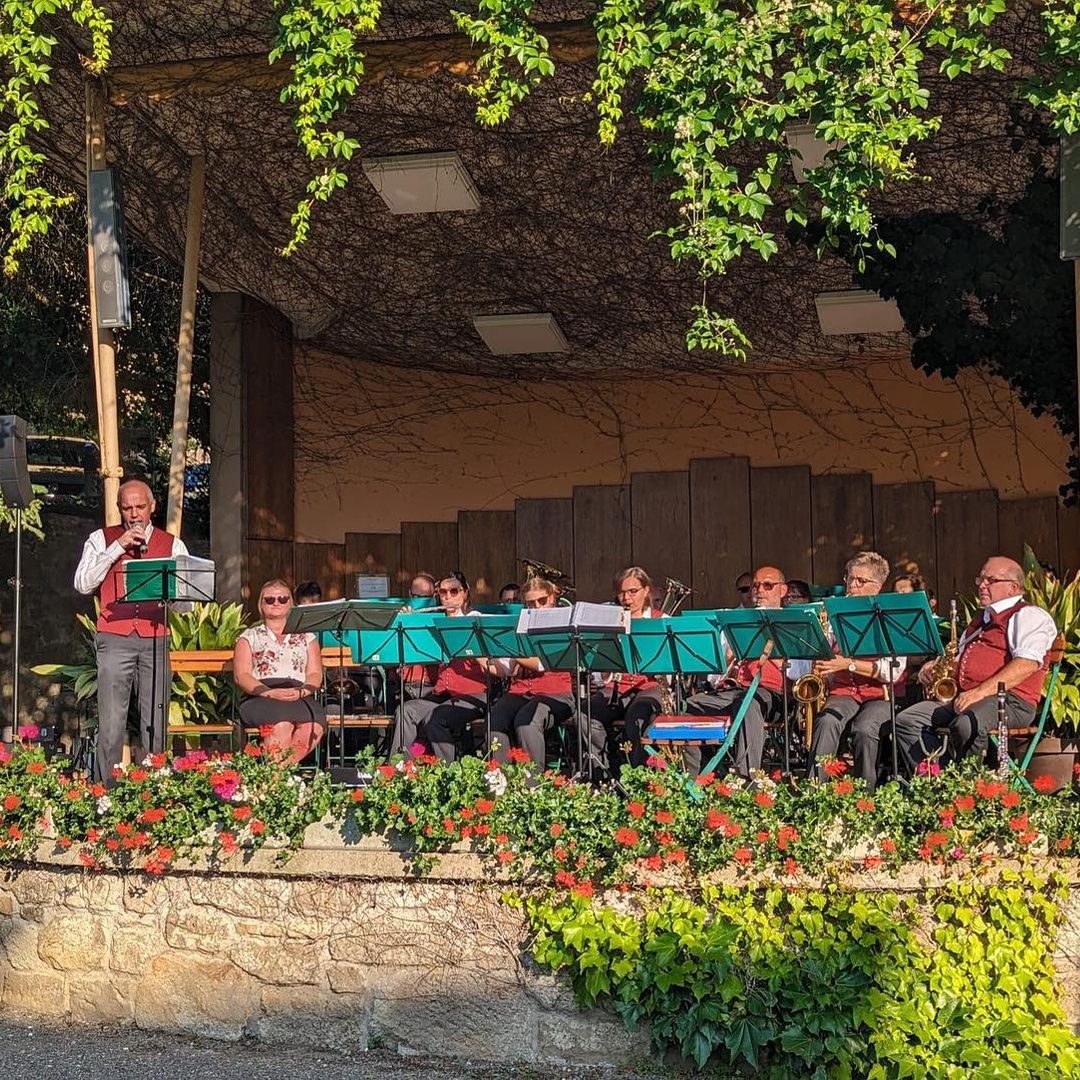 Eine Band spielt auf einer Bühne, die mit roten Blumen und grünen Pflanzen geschmückt ist, unter einem Blätterdach.