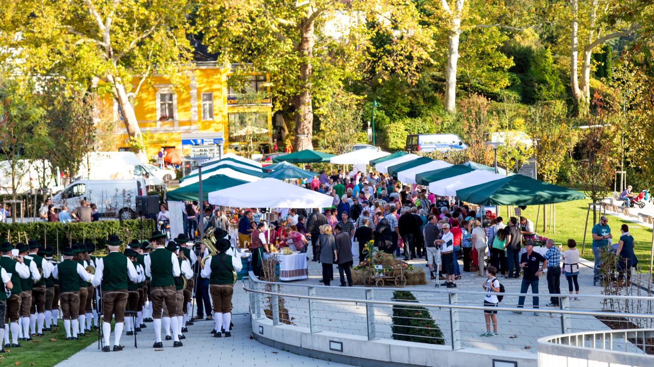 Eine Menschenmenge versammelt sich auf einem Freiluftmarkt mit grünen und weißen Zelten. Musiker in traditioneller Kleidung stehen links, und ein Kind ist rechts. Die Umgebung umfasst ein gelbes Gebäude, Bäume und einen Metallzaun.