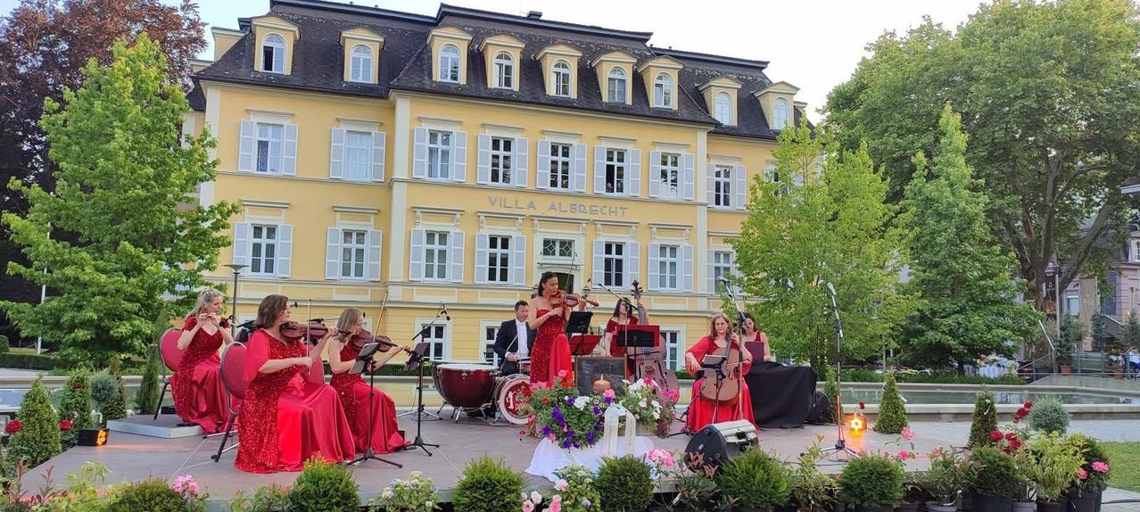 Ein klassisches Musikensemble spielt auf der Bühne vor der Villa Albrecht, mit Musikern, die Violinen, Cello und Schlagzeug spielen, umgeben von Blumen und Grün.