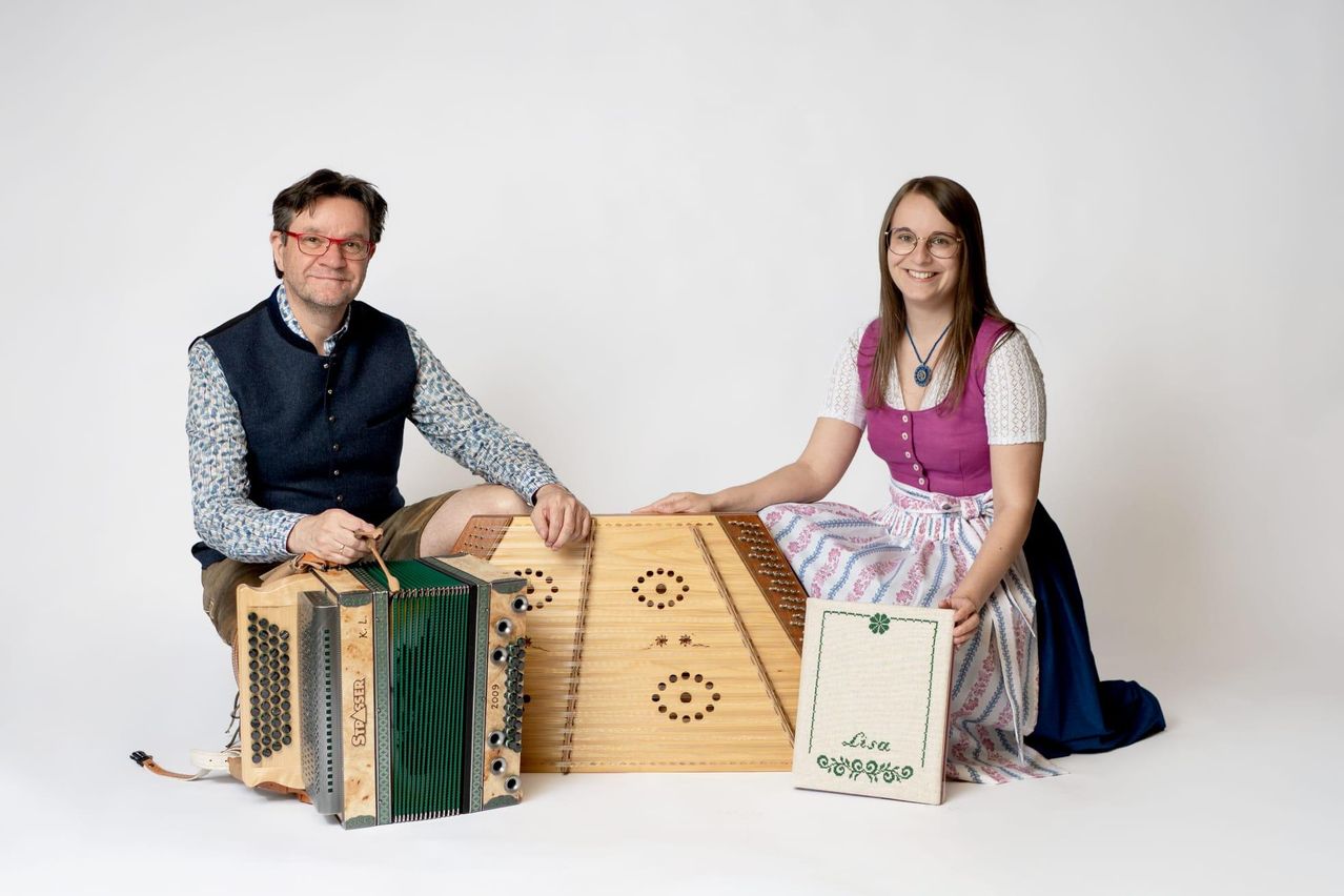 A man and woman sit next to each other with traditional musical instruments. The man holds an accordion and the woman holds a zither. They both wear traditional clothing.