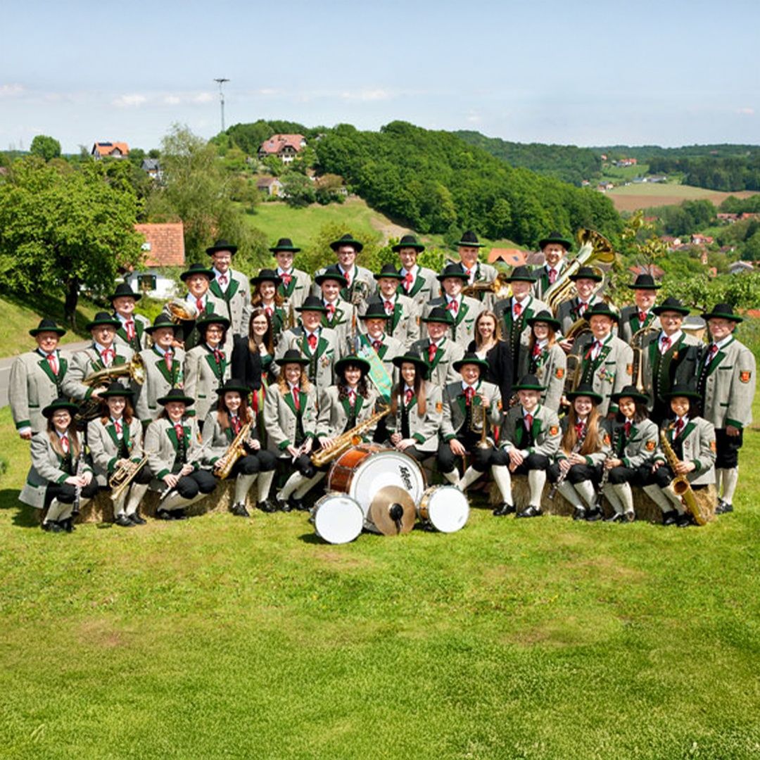 Ein großes Blasorchester in traditioneller deutscher Tracht posiert auf einer Grasfläche mit Instrumenten wie Trompeten und Trommeln vor einer malerischen Kulisse.