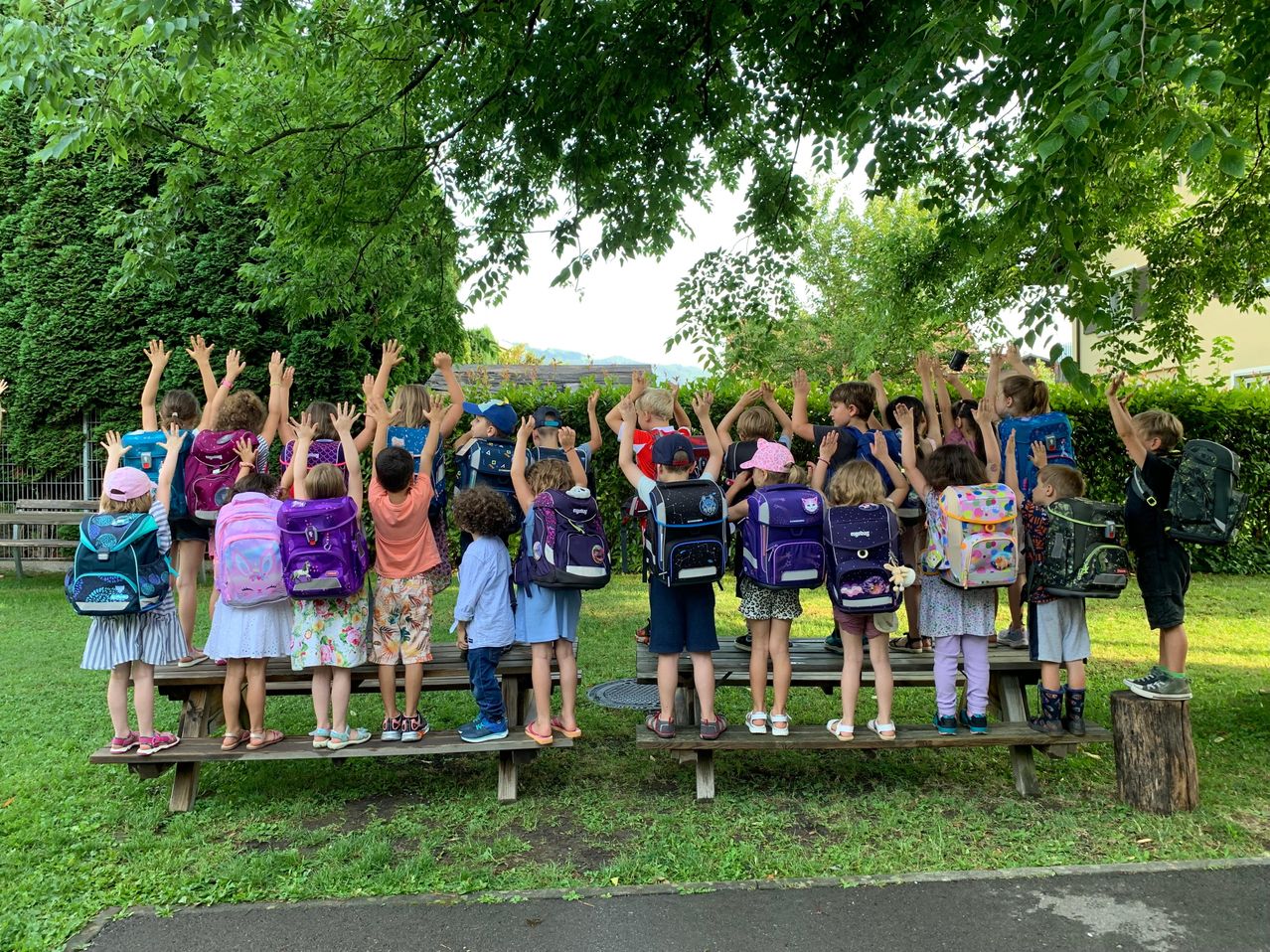 Eine Gruppe von Kindern im Park mit Rucksäcken steht auf einer Bank mit erhobenen Händen. Sie scheinen für ein Foto zu posieren.