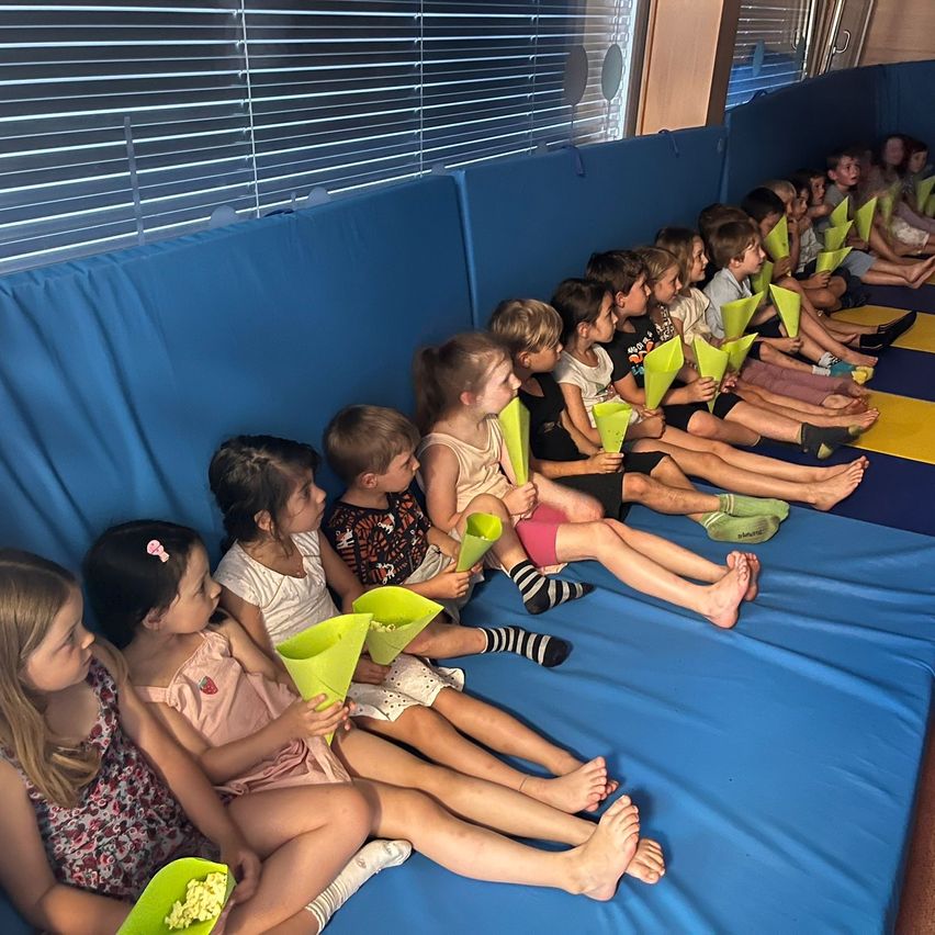 Eine Gruppe von Kindern sitzt auf einem blau gepolsterten Boden, jedes mit einem grünen Kegel in der Hand, wahrscheinlich beim Anschauen eines Films. Sie sind barfuß, einige tragen Socken, und der Raum hat blaue Wände und Jalousien.