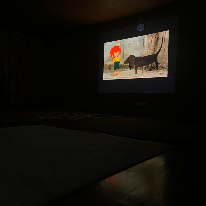 Ein dunkler Raum mit einer Projektionsleinwand, die einen Cartoon von einem Kind und einem Hund zeigt, mit einem Lautsprecher über der Leinwand.