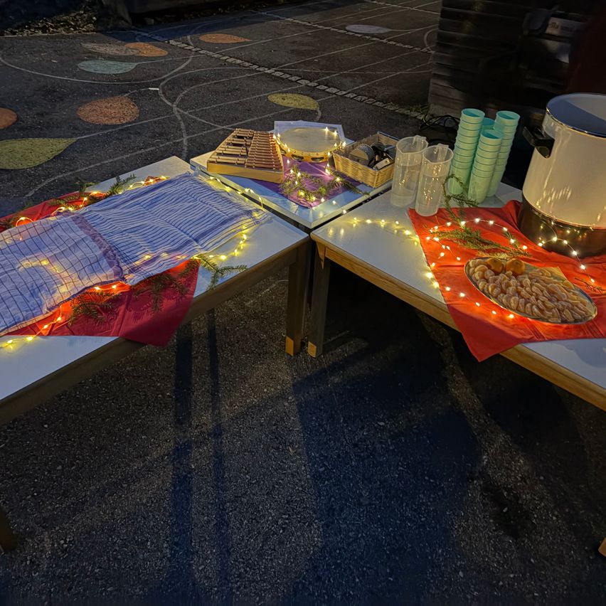 Zwei Tische sind mit festlichen Dekorationen ausgestattet, darunter ein Teller mit Essen, ein Korb und Lichterketten. Ein Tisch hat eine blaue Decke, der andere eine rote. In der Nähe befinden sich Becher und ein Krug.