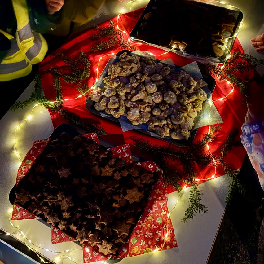 Ein festlicher Tisch mit Tabletts voller dekorierter Kekse, geschmückt mit rotem Tuch und Weihnachtslichtern, mit einer Person im Hintergrund, die eine reflektierende Weste trägt.