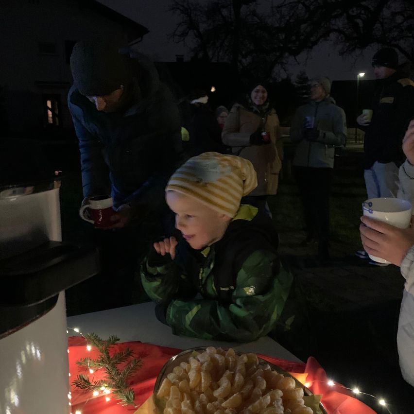 Eine Familie ist nachts draußen versammelt, möglicherweise für eine Feier. Ein Kind sitzt an einem Tisch mit einer Schüssel voller Süßigkeiten vor ihm. Ein Mann steht hinter ihm mit einer Tasse in der Hand. Ein anderer Mann steht im Hintergrund mit einer Tasse in der Hand.