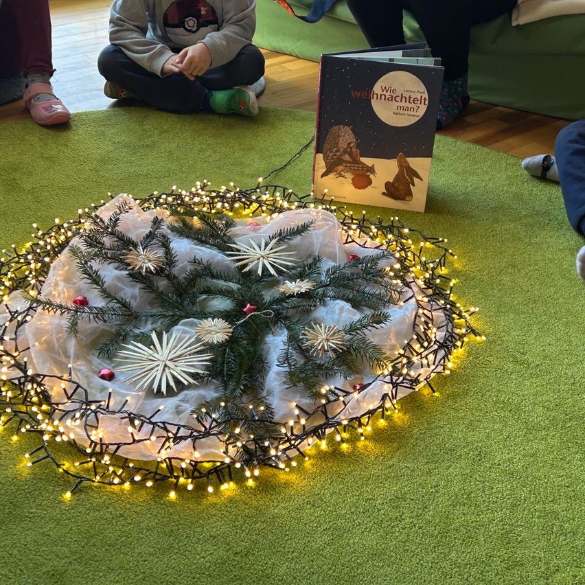 Ein Kind sitzt auf dem Boden neben einem grünen Teppich mit einer Lichtergirlande und einem Weihnachtsbaum. Ein Buch mit dem Titel 'Wie Weihnachten man?' liegt in der Nähe.