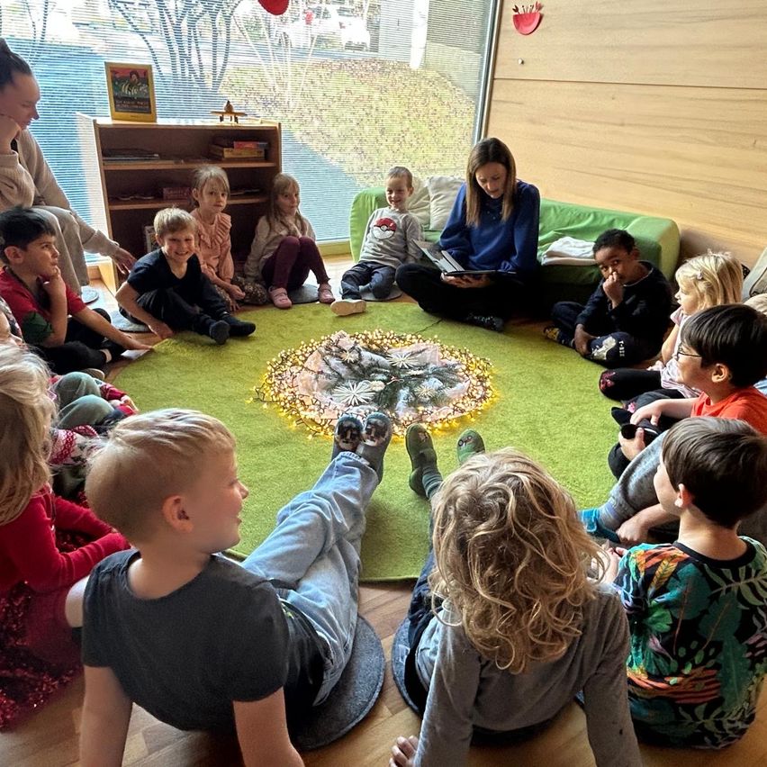 Eine Gruppe von Kindern sitzt im Kreis auf einem grünen Teppich und hört einer Frau zu, die ein Buch liest. Der Raum hat große Fenster mit Blick auf Bäume draußen.