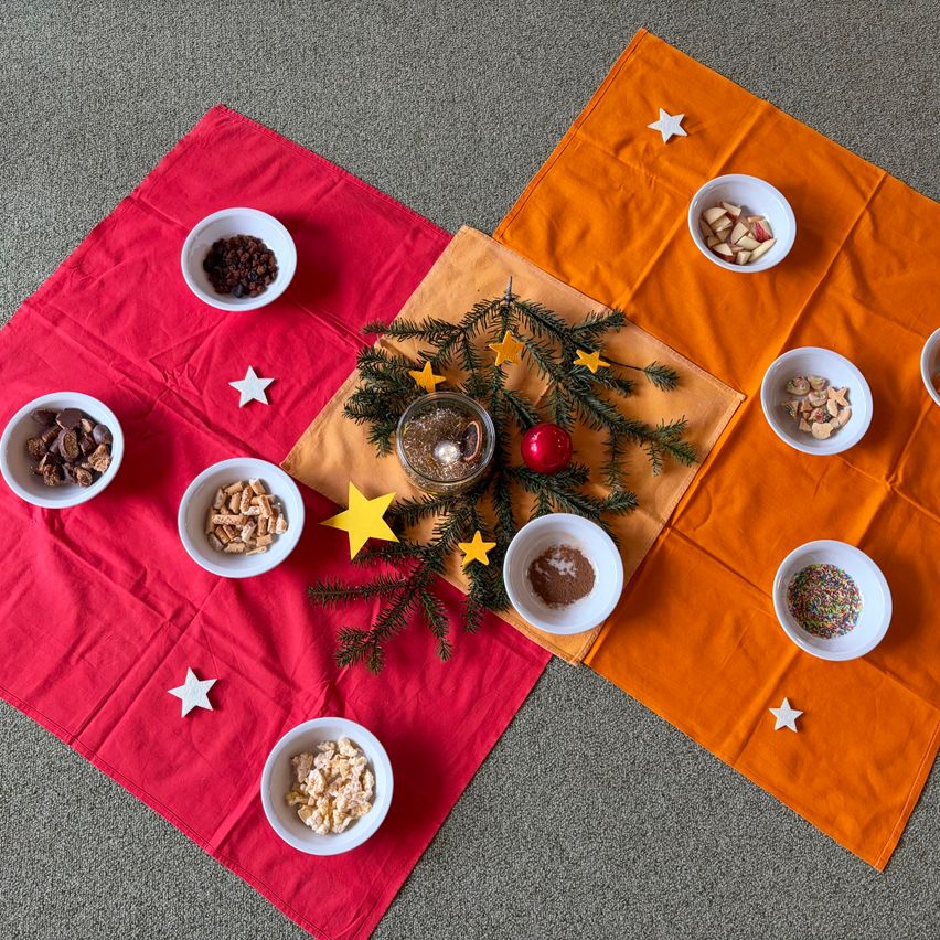 Eine Reihe von Schüsseln mit verschiedenen Snacks und Süßigkeiten ist um einen Weihnachtsbaum auf einem roten und orangefarbenen Tuch angeordnet. Die Schüsseln enthalten Rosinen, Schokolade und Müsli. Der Baum ist mit Sternen und einer roten Kugel geschmückt.