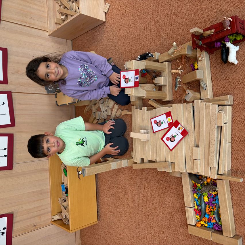 Zwei Kinder sitzen auf dem Boden, lächeln und spielen mit Holzklötzen und Tieren. Der Raum hat eine Holzwand mit Rahmen und einem braunen Teppich.