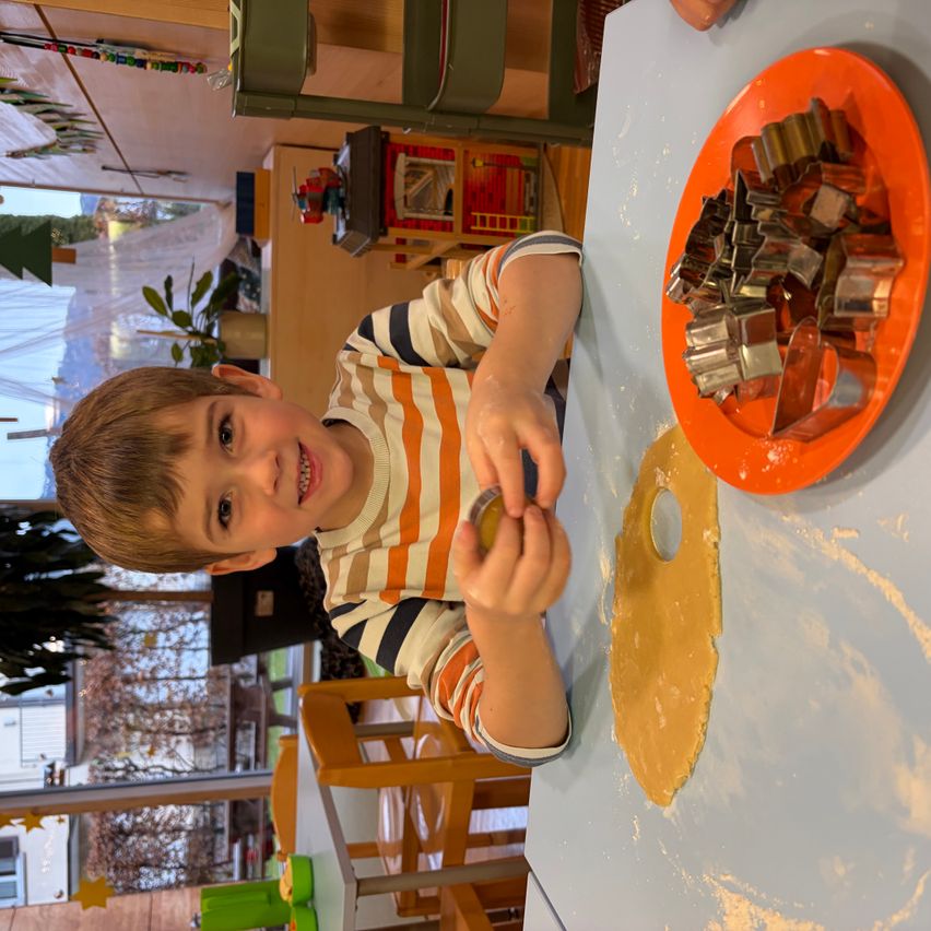 Ein junger Junge benutzt einen Ausstecher auf Teig, mit Ausstechern auf einem Teller zu seiner rechten Seite. Er lächelt und trägt ein gestreiftes Hemd. Hinter ihm zeigt ein Fenster einen Baum und ein Haus.
