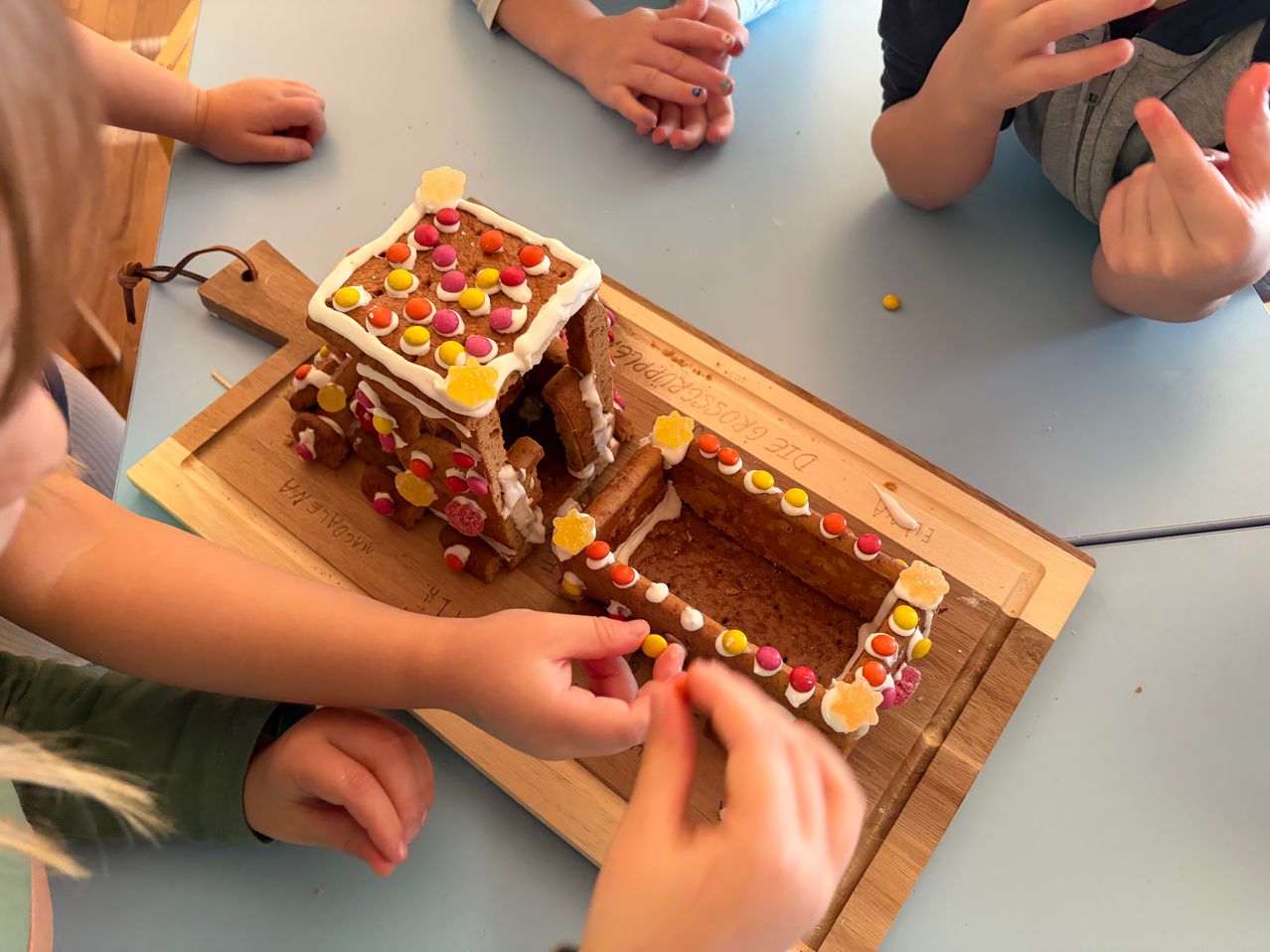 Ein Lebkuchenhaus wird auf einem Holzbrett mit Süßigkeiten verziert und ist von Kindern umgeben.