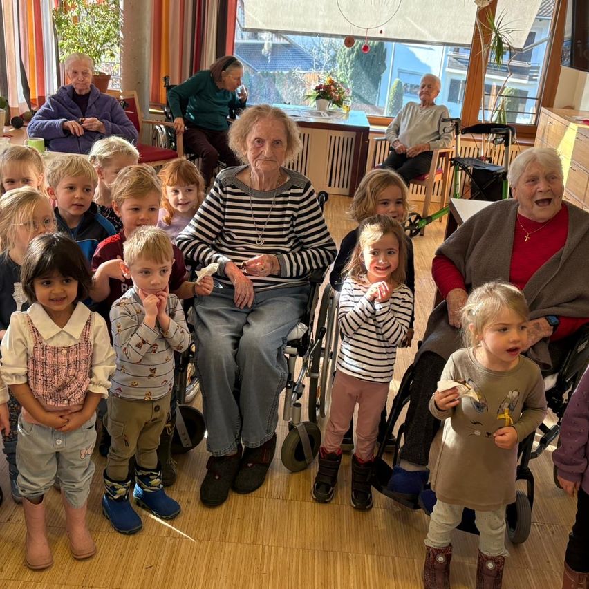 Eine Gruppe älterer Menschen und junger Kinder versammelt sich in einem Raum. Eine ältere Frau sitzt in einem Rollstuhl, mit anderen in der Nähe. Kinder stehen vor ihnen, und ein Kind hält einen Essensgegenstand.