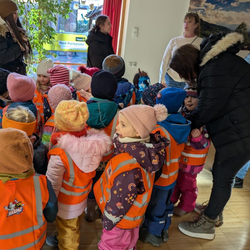 Eine Gruppe von Kindern in orangefarbenen Westen steht in einem Raum und hört einer Erwachsenen zu. Sie tragen Mützen und Winterkleidung. Hinter ihnen stehen Erwachsene und reden. Der Raum hat ein Glasfenster mit einem Schild.