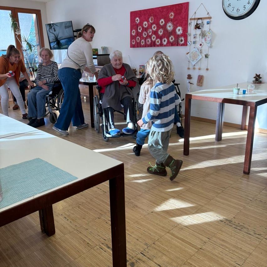 Eine Gruppe von Menschen, darunter eine ältere Frau im Rollstuhl, versammelt sich drinnen. Ein Kind geht auf sie zu. Tische und eine Uhr sind sichtbar.